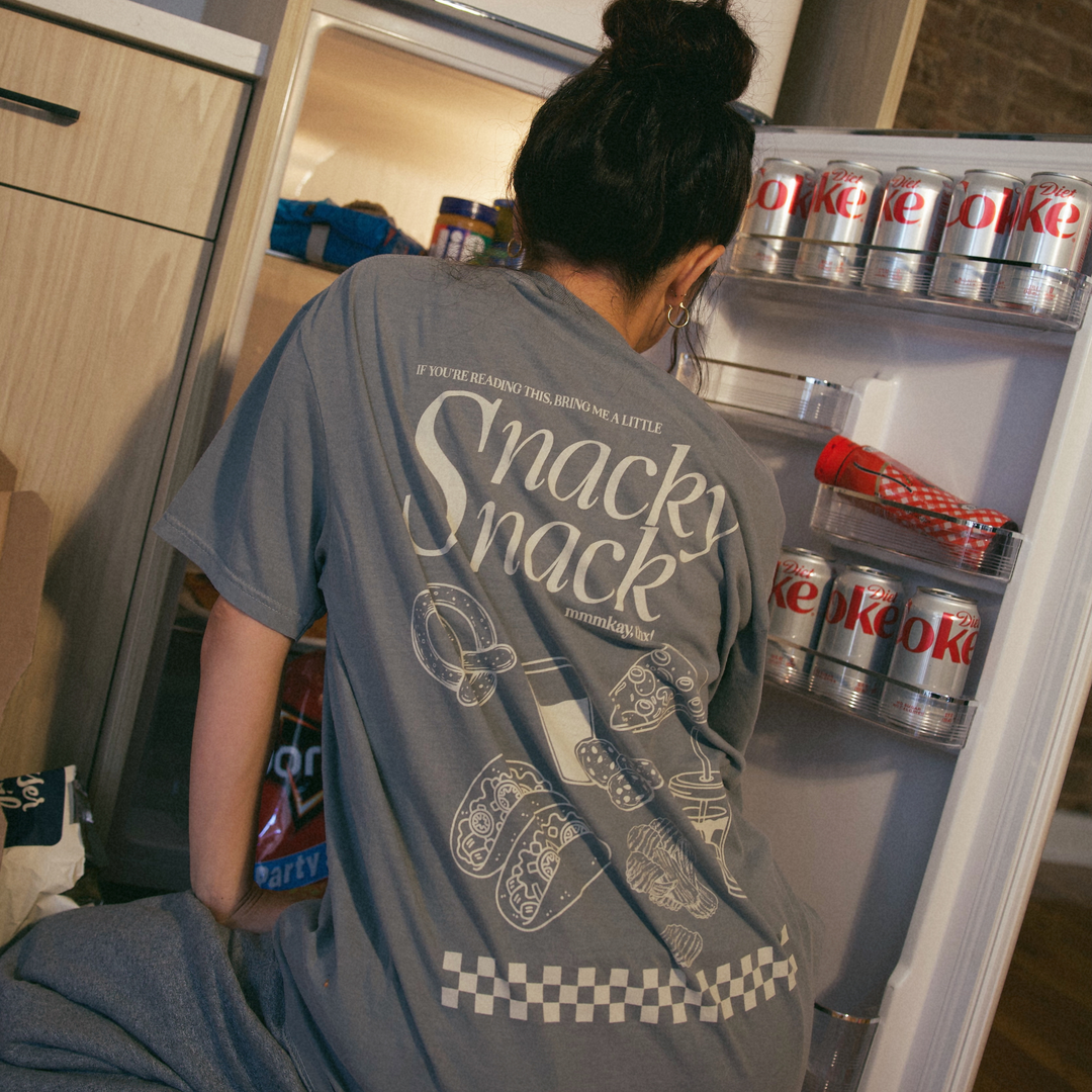 [main] A mom wearing a grey T-shirt with the phrase "if you're reading this bring me a little snacky snack mmmkay" while featuring an assortment of snacks in white font on the back of the T-shirt. 