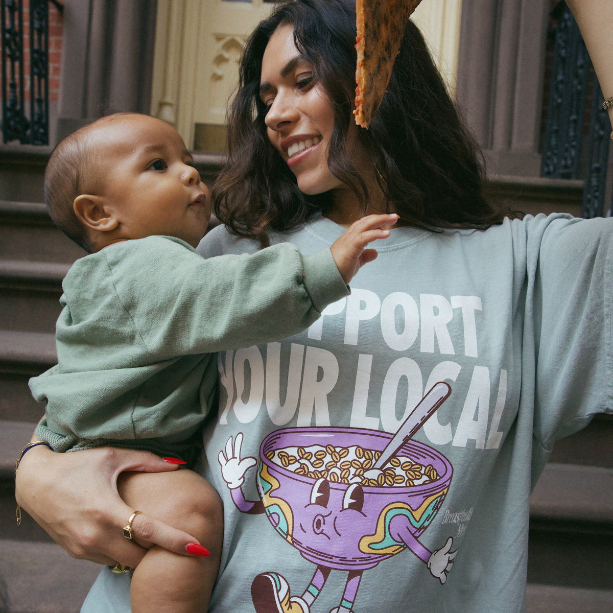 [main] A mom wearing a light sage t-shirt featuring the slogan Support Your Local Breastfeeding Mom and a bright cereal bowl graphic, while feeding her son a piece of pizza.