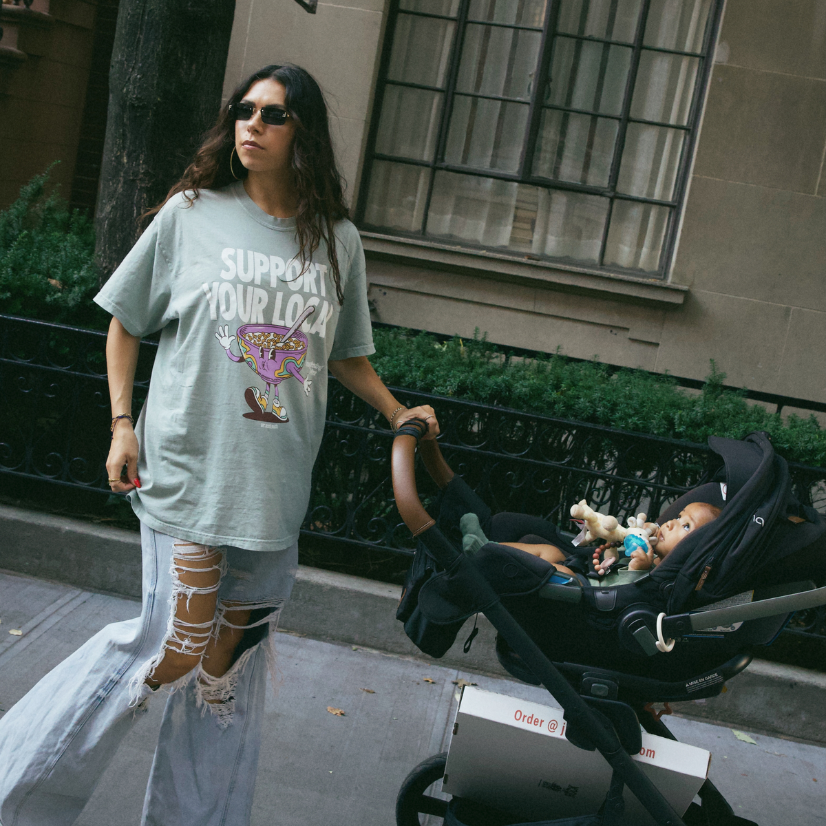 [main] A mom wearing a light sage t-shirt featuring the slogan Support Your Local Breastfeeding Mom and a bright cereal bowl graphic, while walking down the street with a stroller and ripped jeans.