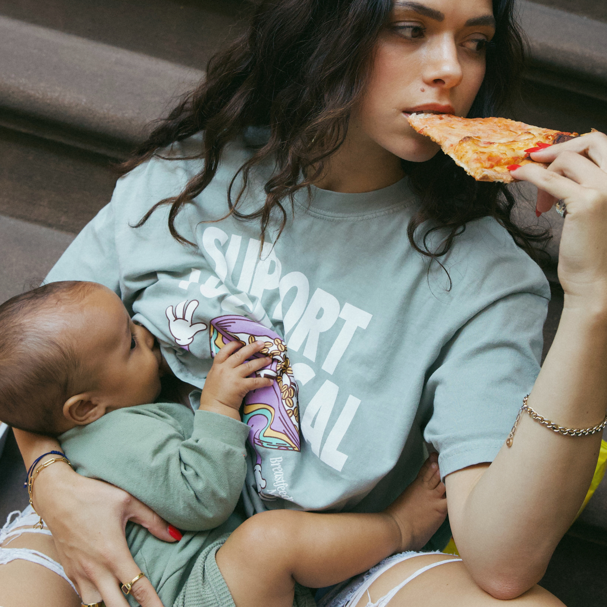 [main] A mom wearing a light sage t-shirt featuring the slogan Support Your Local Breastfeeding Mom and a bright cereal bowl graphic, while eating a piece of pizza and breastfeeding her child.