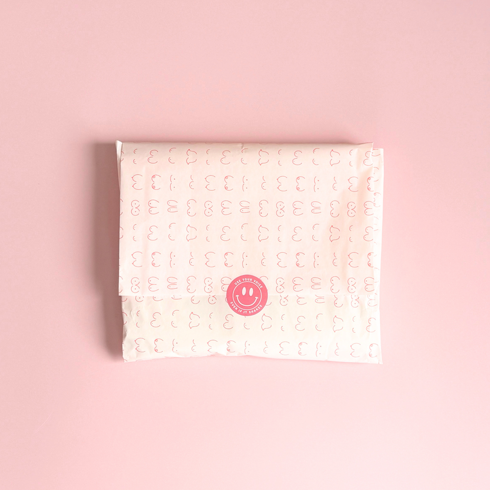 [main] Light Pink tissue paper featuring hot pink shapes of different boobs across the tissue paper to represent breastfeeding, featuring our signature Use Your Voice Smiley Face sticker in the center. 